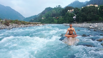 A person meditates in a cross-legged position on a rock in the middle of a flowing river, surrounded by lush green hills and small houses in the background.