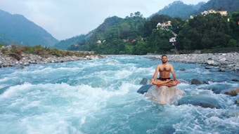 A person meditates in a cross-legged position on a rock in the middle of a flowing river, surrounded by lush green hills and small houses in the background.