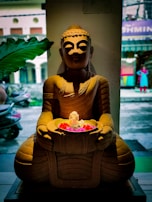 A Buddha statue is placed indoors, holding a bowl filled with flowers. The background shows a street scene slightly out of focus, with a large green leaf prominently featured on the left side.