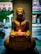 A Buddha statue is placed indoors, holding a bowl filled with flowers. The background shows a street scene slightly out of focus, with a large green leaf prominently featured on the left side.