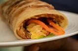 A close-up of a wrap on a plate, featuring filling with sliced vegetables such as tomatoes, lettuce, and possibly grilled chicken. The wrap itself is lightly toasted and has a golden-brown exterior.