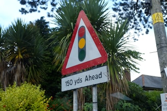 A triangular traffic sign with a red border containing a traffic light symbol, indicating the presence of traffic signals ahead. The sign is mounted on a pole and accompanied by a rectangular sign stating '150 yds Ahead'. In the background, there are tall palm trees and lush green foliage, with a glimpse of a residential building. A utility pole with a warning sign is also visible.
