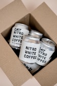 A cardboard box containing several cans of Nitro White Coffee and Oat Nitro White Coffee. The cans are neatly arranged inside the box, featuring bold black text labeling against a light-colored background.