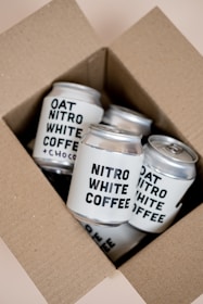 A cardboard box containing several cans of Nitro White Coffee and Oat Nitro White Coffee. The cans are neatly arranged inside the box, featuring bold black text labeling against a light-colored background.