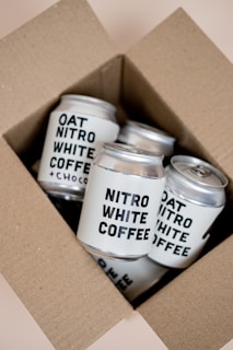 A cardboard box containing several cans of Nitro White Coffee and Oat Nitro White Coffee. The cans are neatly arranged inside the box, featuring bold black text labeling against a light-colored background.