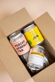A cardboard box contains several colorful canned beverages. The visible cans feature various labels with bold text, including 'Peachy Sparkling Tea', 'Xun Jun Xun', and 'Pushers Def Natural Immunity Booster'. The box is open, revealing the neatly arranged cans inside.