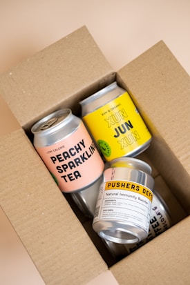 A cardboard box contains several colorful canned beverages. The visible cans feature various labels with bold text, including 'Peachy Sparkling Tea', 'Xun Jun Xun', and 'Pushers Def Natural Immunity Booster'. The box is open, revealing the neatly arranged cans inside.
