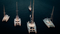 Several sailboats and catamarans are positioned on calm, dark blue water. The boats are arranged in an organized pattern, illuminated by soft lighting that highlights their white hulls against the deep color of the ocean.