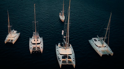 Several sailboats and catamarans are positioned on calm, dark blue water. The boats are arranged in an organized pattern, illuminated by soft lighting that highlights their white hulls against the deep color of the ocean.