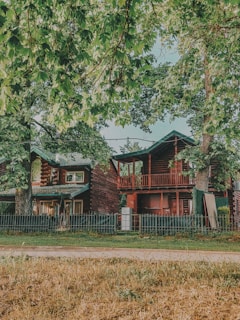 A rustic wooden house surrounded by large green trees and a small front yard with grass. The house has two stories, a front porch, and a balcony. A picket fence runs along the front of the property.