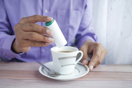 A person in a purple shirt is adding sweetener to a cup of tea, which is placed on a saucer with a teaspoon on a wooden table. The setting appears to be indoors with a soft, light curtain in the background.