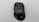 A black gaming mouse with multiple buttons and a silver scroll wheel, labeled G700s. The surface has a sleek, ergonomic design and distinct textured patterns.