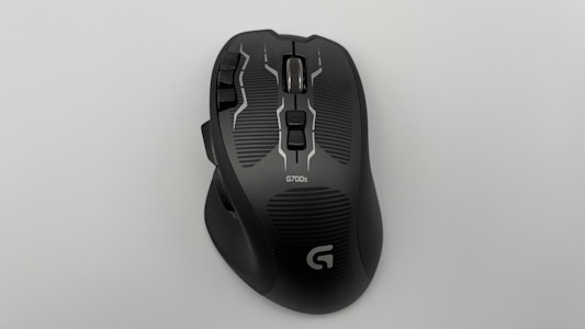 A black gaming mouse with multiple buttons and a silver scroll wheel, labeled G700s. The surface has a sleek, ergonomic design and distinct textured patterns.
