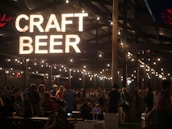 A lively evening setting at a craft beer event with a large illuminated sign reading 'Craft Beer' hanging above. The area is bustling with people socializing under a wooden structure decorated with string lights, creating a warm and inviting atmosphere.