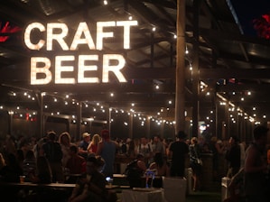 A lively evening setting at a craft beer event with a large illuminated sign reading 'Craft Beer' hanging above. The area is bustling with people socializing under a wooden structure decorated with string lights, creating a warm and inviting atmosphere.