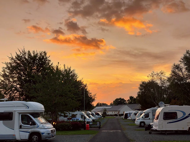 A serene campsite scene at sunset, with several caravans and motorhomes parked on either side of a central pathway. The sky is ablaze with vibrant orange and yellow hues, contrasted against a few dark clouds. Lush green trees frame the campsite, enhancing the natural and tranquil setting.