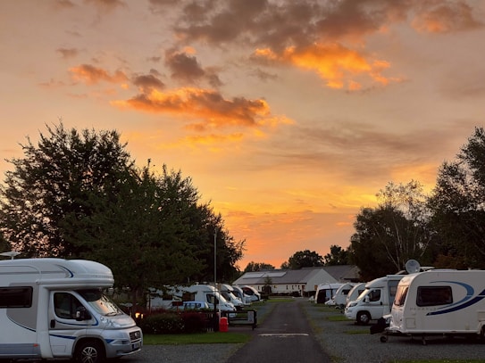 A serene campsite scene at sunset, with several caravans and motorhomes parked on either side of a central pathway. The sky is ablaze with vibrant orange and yellow hues, contrasted against a few dark clouds. Lush green trees frame the campsite, enhancing the natural and tranquil setting.