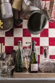 A cozy kitchen scene with a collection of kitchen utensils and cookware hanging on the wall above a tiled backsplash. On the counter, there are two bottles; one is a green glass bottle and the other is a champagne bottle alongside a glass partially filled with champagne. There are various glasses arranged neatly along with a set of utensils in a holder. A dark hat is casually placed among the hanging pots and pans.