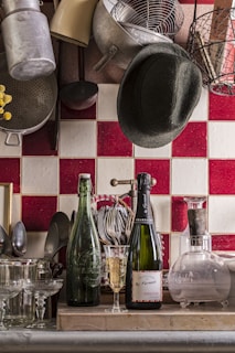 A cozy kitchen scene with a collection of kitchen utensils and cookware hanging on the wall above a tiled backsplash. On the counter, there are two bottles; one is a green glass bottle and the other is a champagne bottle alongside a glass partially filled with champagne. There are various glasses arranged neatly along with a set of utensils in a holder. A dark hat is casually placed among the hanging pots and pans.