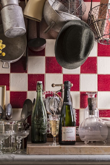 A cozy kitchen scene with a collection of kitchen utensils and cookware hanging on the wall above a tiled backsplash. On the counter, there are two bottles; one is a green glass bottle and the other is a champagne bottle alongside a glass partially filled with champagne. There are various glasses arranged neatly along with a set of utensils in a holder. A dark hat is casually placed among the hanging pots and pans.