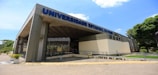 A large, modern building with a wide entrance and the name 'Universidade Estadual do Piauí' prominently displayed in blue letters on the facade. The structure features a contemporary design with a mix of concrete and glass. There are several potted plants lining the walkway leading to the entrance. The sky is bright blue with a few scattered clouds.