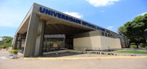 A large, modern building with a wide entrance and the name 'Universidade Estadual do Piau&iacute;' prominently displayed in blue letters on the facade. The structure features a contemporary design with a mix of concrete and glass. There are several potted plants lining the walkway leading to the entrance. The sky is bright blue with a few scattered clouds.