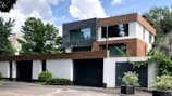 A modern two-story house with a flat roof, featuring a mix of white, gray, and brown exteriors. Large glass windows are prominent, allowing for natural light. The house is surrounded by lush greenery, including bushes and trees, against a partly cloudy sky.