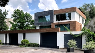 A modern two-story house with a flat roof, featuring a mix of white, gray, and brown exteriors. Large glass windows are prominent, allowing for natural light. The house is surrounded by lush greenery, including bushes and trees, against a partly cloudy sky.