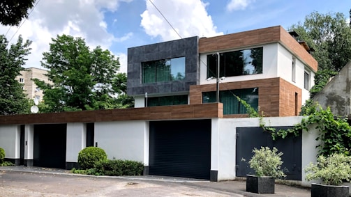 A modern two-story house with a flat roof, featuring a mix of white, gray, and brown exteriors. Large glass windows are prominent, allowing for natural light. The house is surrounded by lush greenery, including bushes and trees, against a partly cloudy sky.