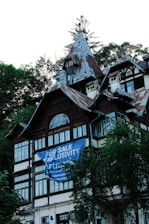 A multi-story house with intricate architectural details, featuring steep, pointed roofs and multiple windows. A large blue banner with white text 'For Sale Exclusivity' partially covers the front of the building. The structure is surrounded by lush green trees.