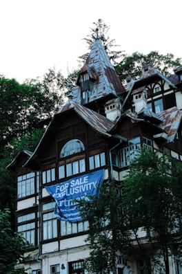 A multi-story house with intricate architectural details, featuring steep, pointed roofs and multiple windows. A large blue banner with white text 'For Sale Exclusivity' partially covers the front of the building. The structure is surrounded by lush green trees.
