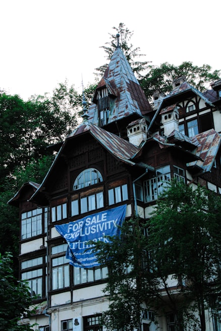 A multi-story house with intricate architectural details, featuring steep, pointed roofs and multiple windows. A large blue banner with white text 'For Sale Exclusivity' partially covers the front of the building. The structure is surrounded by lush green trees.