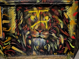 A vibrant graffiti artwork of a lion's face covers a wooden surface. The lion's eyes shine with bright yellow and its mane is an eclectic mix of vivid reds, oranges, yellows, and black. The dynamic strokes and colorful patterns create an intense and energetic visual impact.