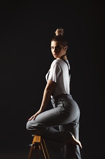 An editorial-style photo of a model in a flowing off-white shirt and tailored pants, seated on a minimalist wooden chair with soft natural light.