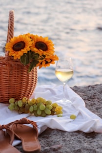yellow sunflower in brown woven basket beside clear wine glass on white textile