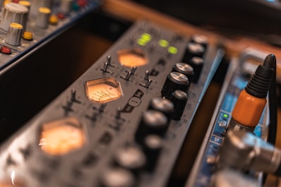 A close-up view of audio equipment featuring various control knobs and LED indicators. The foreground shows a VU meter with glowing orange backlighting, and to the side, there are connectors for audio cables, including an orange plug.
