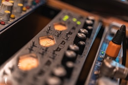 A close-up view of audio equipment featuring various control knobs and LED indicators. The foreground shows a VU meter with glowing orange backlighting, and to the side, there are connectors for audio cables, including an orange plug.