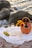 A picturesque picnic setup by the seashore features two glasses of white wine, an open book, a bunch of green grapes, and a wicker basket filled with sunflowers and a wine bottle, all placed on a white cloth. Gentle waves and large rocks create a serene backdrop.
