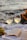 Two glasses of white wine are placed on a white cloth beside an open book. Green grapes are on the left, and orange flowers are on the right. The background features a blurred view of water, suggesting a scenic or beach setting.