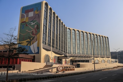 A modern building with an artistic mural on one side. The mural depicts a stylized human figure interacting with abstract shapes and elements. The building features a series of tall, arched windows giving it an elegant appearance. The scene is set in daylight under a clear blue sky, and there is a tree and a clear road in the foreground.