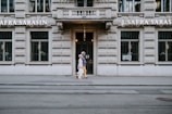 A pair of people walking in front of an ornate stone building with barred windows. The facade features the name 'J. Safra Sarasin'. The street is empty with visible tram tracks running parallel to the building.