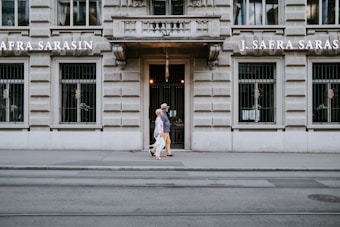 A pair of people walking in front of an ornate stone building with barred windows. The facade features the name 'J. Safra Sarasin'. The street is empty with visible tram tracks running parallel to the building.