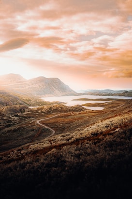 A serene landscape featuring rolling hills and a winding road leading through the terrain. The sky is filled with dramatic clouds and warm tones, casting an enchanting glow over the scene. In the background, mountains rise gently from the earth, and a body of water reflects the hues of the sky.