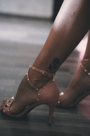 A pair of legs wearing pink high-heeled shoes with ankle straps and studded embellishments is shown. There is a tattoo of a floral design on one of the legs. The floor is a dark, polished wood and the lighting is soft, creating a slightly muted ambiance.