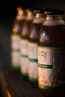 A row of glass bottles containing a beverage, with visible labels indicating they are organic tea. The labels are green and white, and the bottles are placed on a wooden surface with a blurred background.