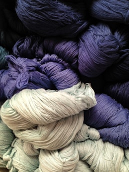 A collection of colorful yarn skeins arranged in layers, showcasing deep blue, dark purple, and light grey hues. The yarns have a soft, textured appearance, highlighting the intricate fibers and subtle sheen.