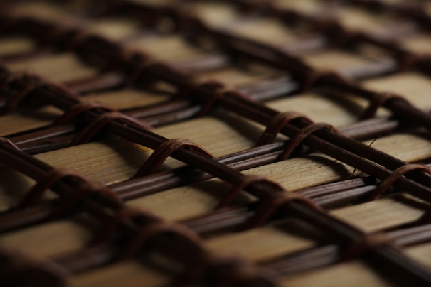 A close-up view of a woven pattern made from intersecting brown strands, possibly a basket or mat. The strands create a grid-like design with visible texture and shadows.