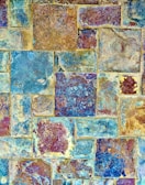 A textured mosaic of stone tiles displaying a variety of shapes and sizes. The tiles have a weathered appearance with a mixture of earthy and blue tones, featuring intricate patterns and natural formations.