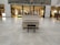 A white upright piano is placed in a spacious shopping mall with shiny marble flooring. There are retail stores in the background with various products displayed in well-lit shelves. A black piano bench is positioned in front of the piano.