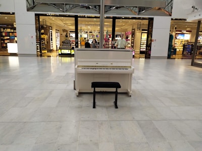 A white upright piano is placed in a spacious shopping mall with shiny marble flooring. There are retail stores in the background with various products displayed in well-lit shelves. A black piano bench is positioned in front of the piano.